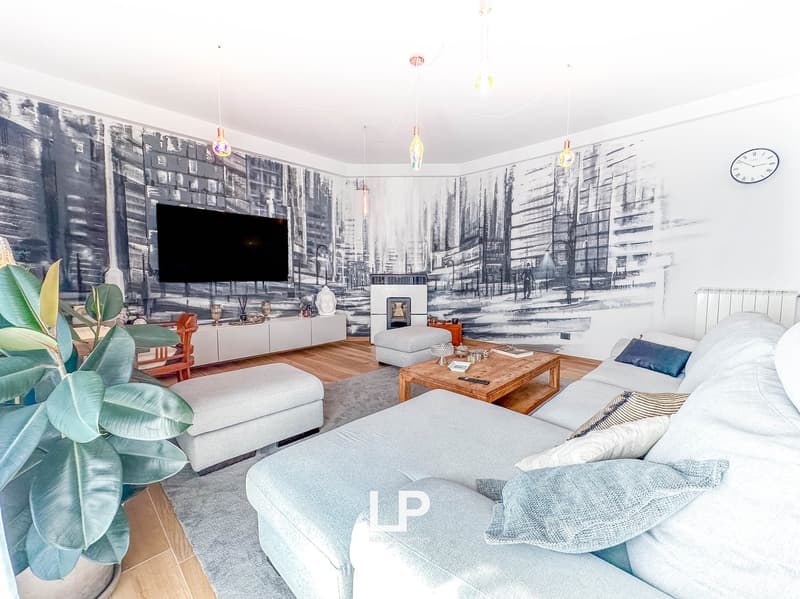 Large living room with a city skyline mural on the walls, hardwood floors, gray sofas, wooden coffee table, and decorative lighting fixtures. There is a fireplace and a balcony or terrace visible.