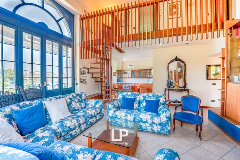 Spacious living room with large windows, blue and white floral patterned sofas, wooden staircase, glass coffee table, and a decorative mirror on the wall