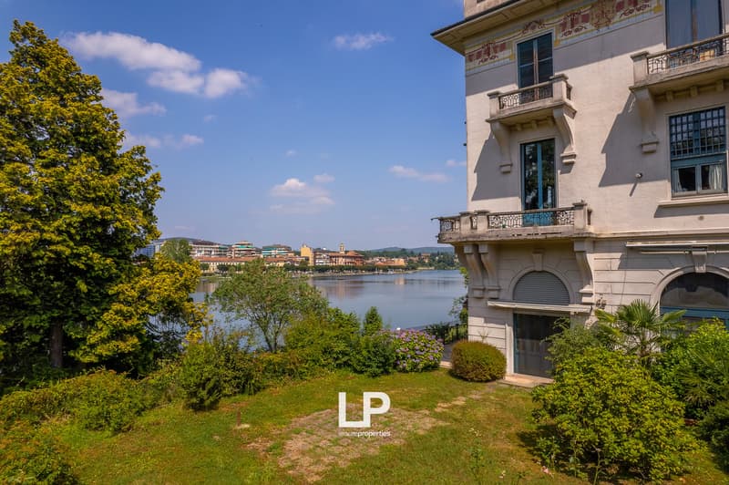 Ornate historic building with balconies overlooking a lake, surrounded by lush greenery and trees