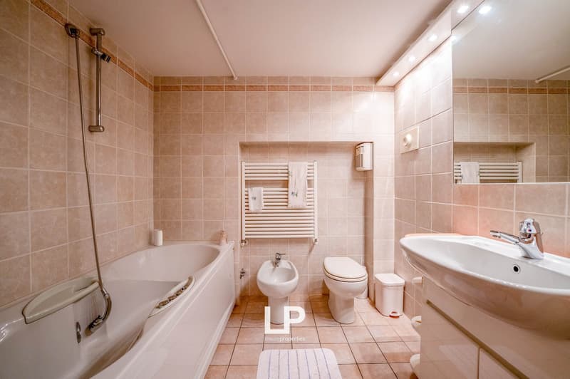 Tiled bathroom with bathtub, toilet, sink, and towel rack. The bathroom appears to be well-lit with a bright and clean design.