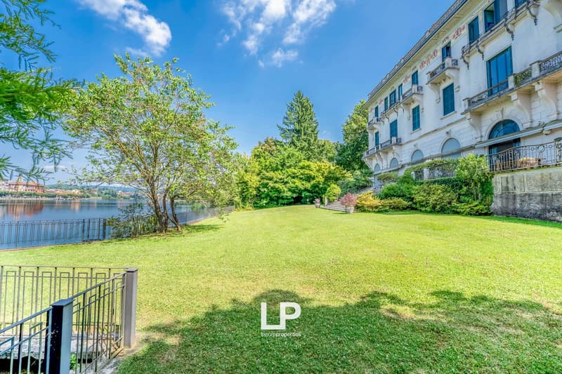 The image shows a large, multi-story apartment building surrounded by a well-manicured lawn and lush greenery. The building has a classic, elegant architectural style with balconies and ornate details. The lawn in front of the building is expansive and we