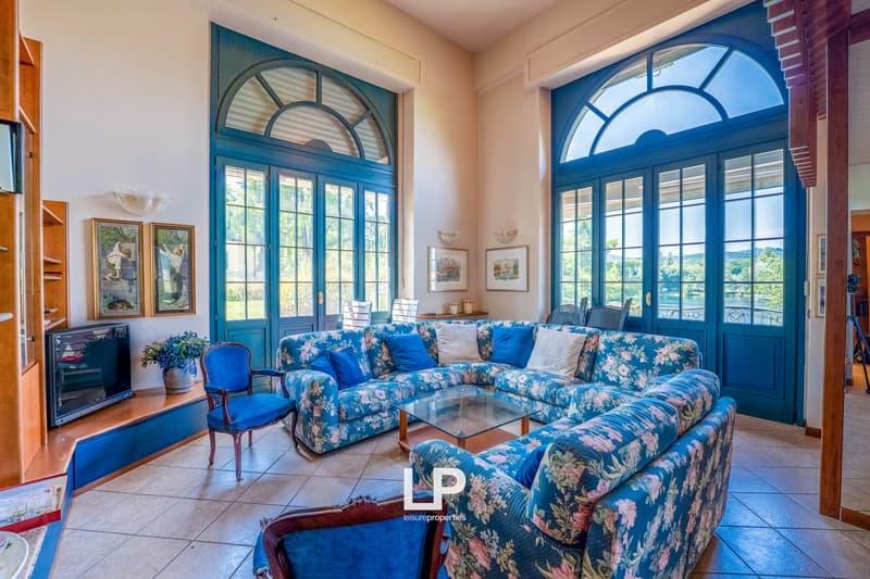 Spacious living room with large arched windows providing a scenic view, blue floral patterned sofa set, glass coffee table, and framed artwork on the walls