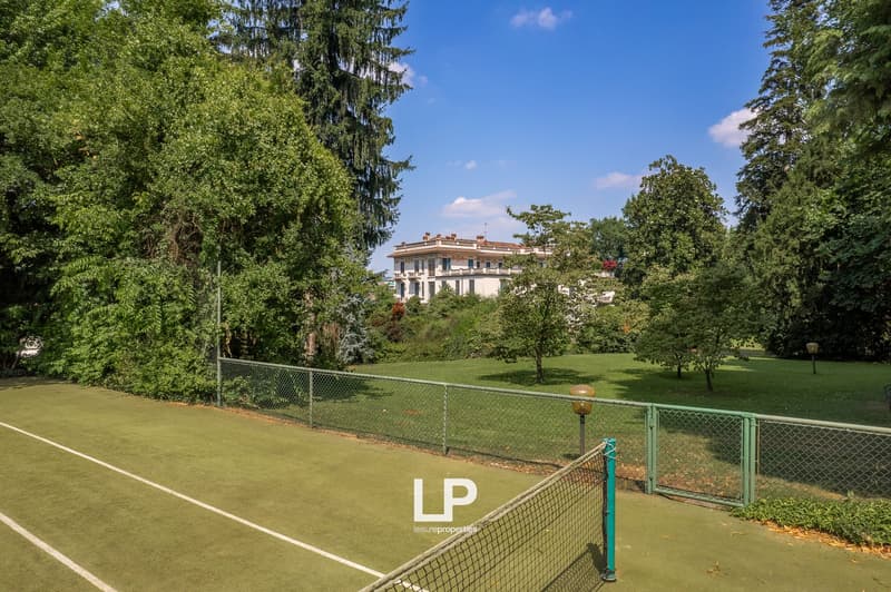 Tennis court surrounded by lush greenery, including tall trees and a well-manicured lawn. A large multi-story building is visible in the background, suggesting this is part of a larger property or estate.