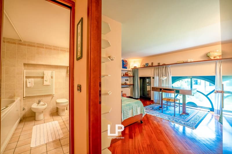 Open-plan studio apartment with hardwood floors, large windows, and a balcony. The bathroom is visible through the open doorway, with tiled floors and walls. The living and sleeping areas are combined in the main room, which has built-in shelving and a sm