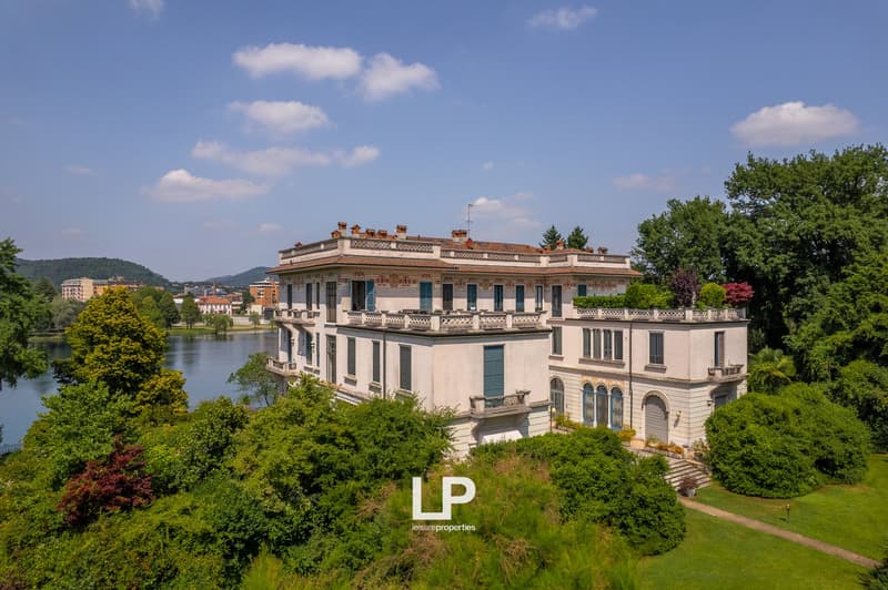 Large, multi-story historic villa or palace with ornate architectural details, surrounded by lush greenery and overlooking a body of water. The building has a grand, classical style with balconies, arched windows, and decorative elements on the facade.