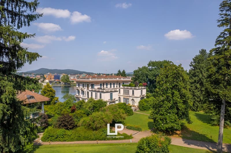 A large, multi-story building with a classical architectural style, surrounded by lush greenery and a well-manicured garden. The building appears to be situated on a hill, offering a scenic view of the surrounding landscape, which includes mountains and a