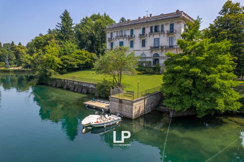 A historic multi-story building with balconies, surrounded by lush greenery and situated next to a calm, turquoise-colored body of water. There is a small boat docked in the water in front of the building.