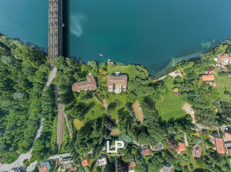 This image shows an aerial view of a scenic landscape with a large body of water, surrounded by lush green forests and buildings. There is a tall structure, possibly a tower or bridge, visible in the center of the image. The overall scene appears to be a