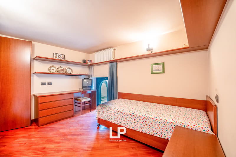 Spacious bedroom with wooden furniture, including a dresser and shelves. The room has a double bed with a colorful patterned bedspread. There is also a small TV and a refrigerator visible.