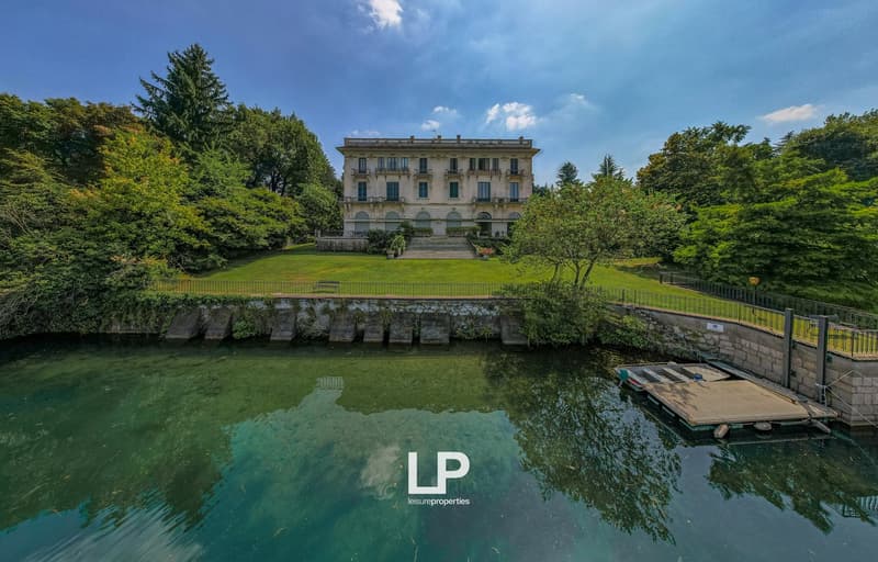 Large historic villa with a classical architectural style, surrounded by lush greenery and a pond. The building has multiple floors and balconies, and is situated in a scenic, park-like setting.