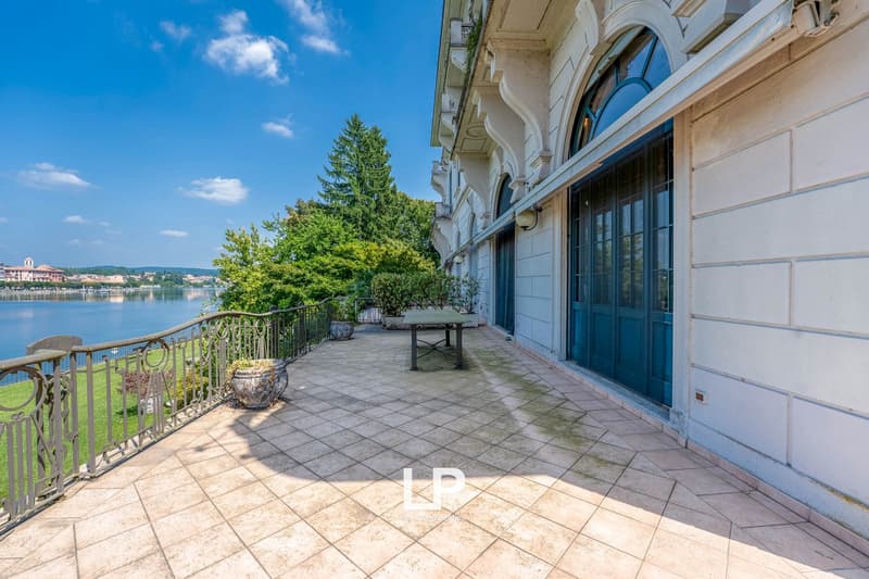Ornate, historic building with arched windows and a balcony overlooking a lake or river. The building has a stone facade and is surrounded by lush greenery. There is a tiled patio area with a table and chairs on the balcony.