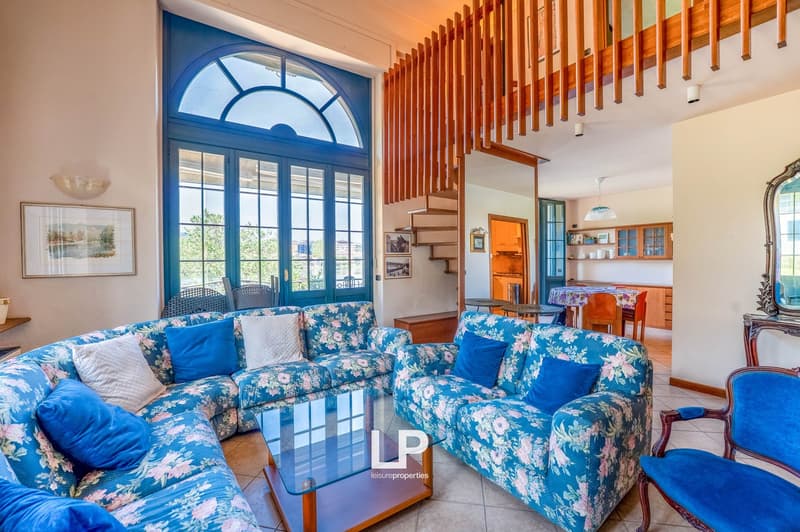 Spacious living room with large arched window, blue and white floral patterned sofas, wooden staircase, and open kitchen area