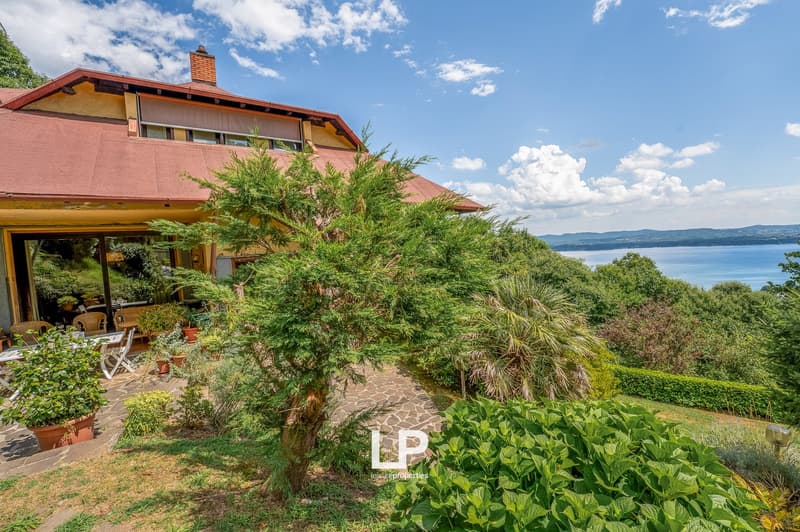 Two-story house with red roof, surrounded by lush greenery and vegetation, overlooking a body of water, with a patio or balcony area visible