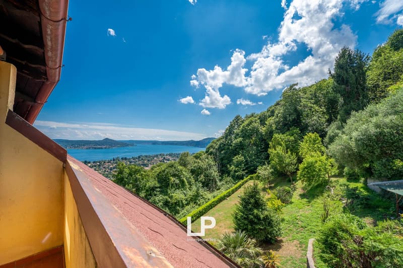 The image shows a scenic view of a lakeside town surrounded by lush green forests and hills. The sky is clear with fluffy white clouds, and the lake in the distance appears calm and serene. The foreground includes a tiled roof and some vegetation, suggest