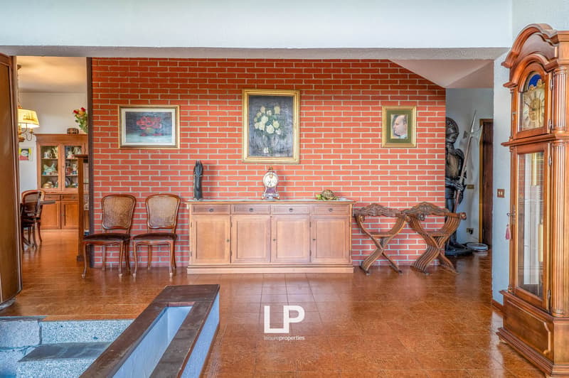 Spacious living room with brick walls, hardwood floors, antique furniture including a wooden cabinet, chairs, and a grandfather clock. The room is decorated with framed artwork on the walls.