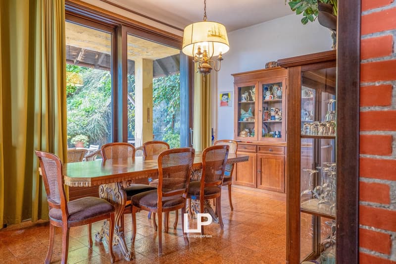 Spacious dining room with large windows overlooking a lush garden, wooden dining table and chairs, a wooden cabinet displaying decorative items, and a chandelier lighting up the room.
