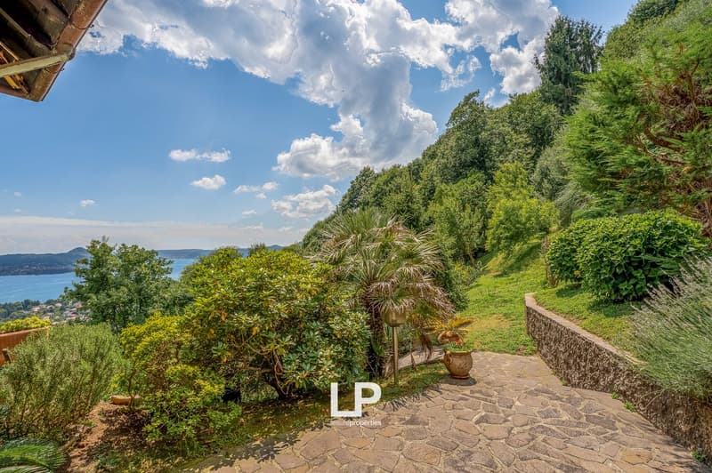 Lush, green garden with various plants and trees, stone pathway, overlooking a scenic view of the water and mountains in the distance
