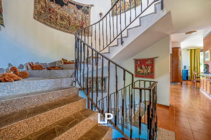 Ornate tapestry on the wall, stone staircase with wrought iron railing, wooden furniture, and a fireplace visible in the background.