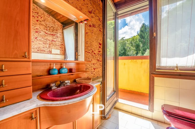 Spacious bathroom with a large red oval-shaped sink, wooden cabinets, and a balcony with a view of the surrounding greenery