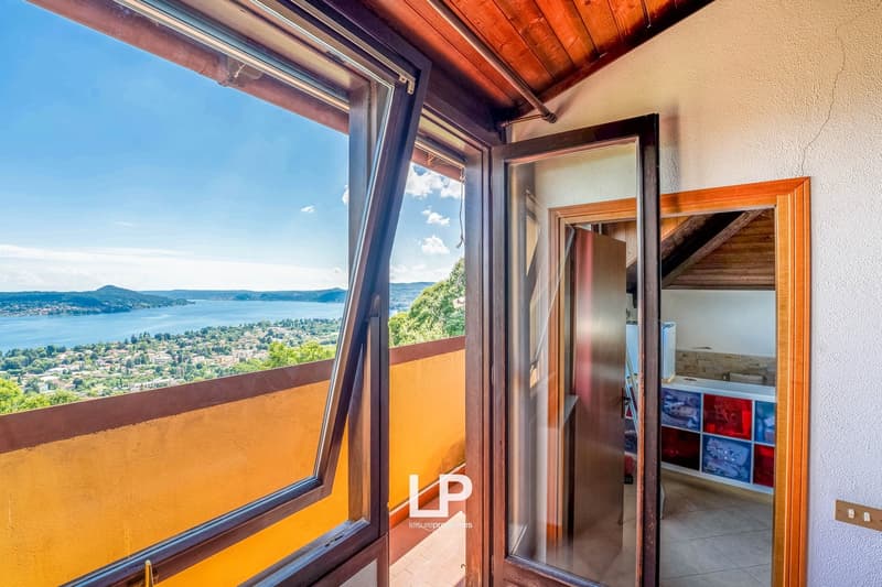 This image shows a room with large windows offering a scenic view of a lake and surrounding landscape. The room has wooden beams on the ceiling and an open doorway leading to another area. The walls are painted in warm, earthy tones, creating a cozy and i