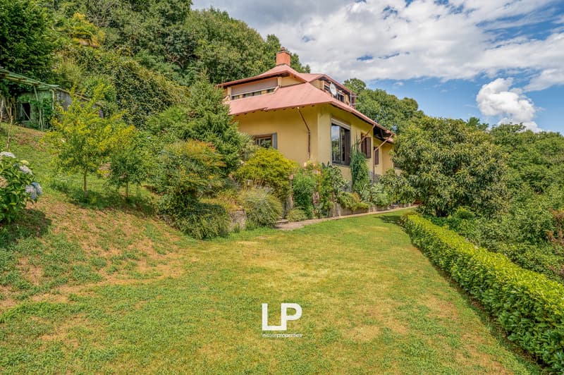 2-story house with a red roof, surrounded by lush greenery and a well-maintained garden. The house has large windows and a balcony or terrace overlooking the scenic landscape.
