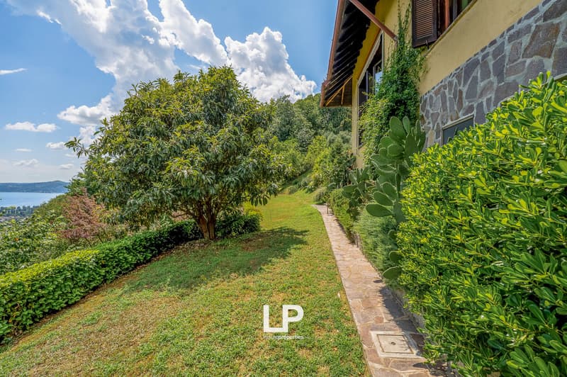 Two-story house with stone exterior, surrounded by lush greenery including trees, shrubs, and a paved walkway. The property offers a scenic view of the surrounding landscape.