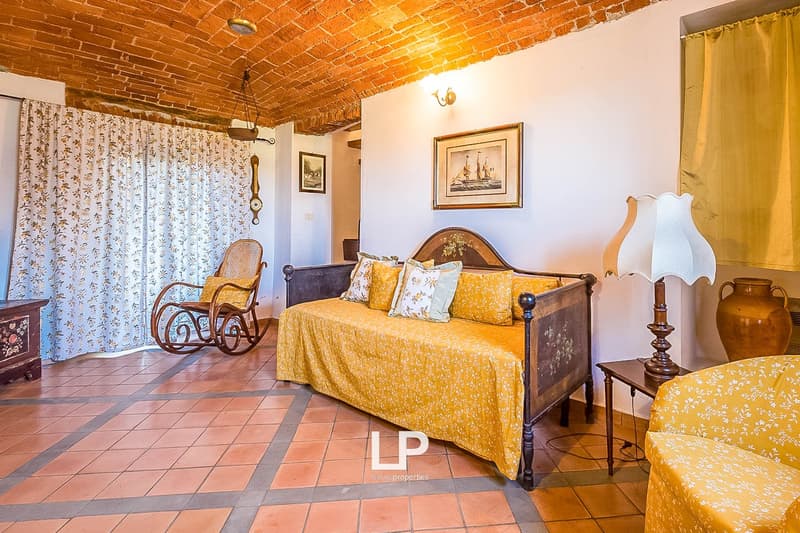 Cozy bedroom with exposed brick ceiling, wooden furniture, floral curtains, and a fireplace. The room has a warm, rustic feel with terracotta tile flooring.