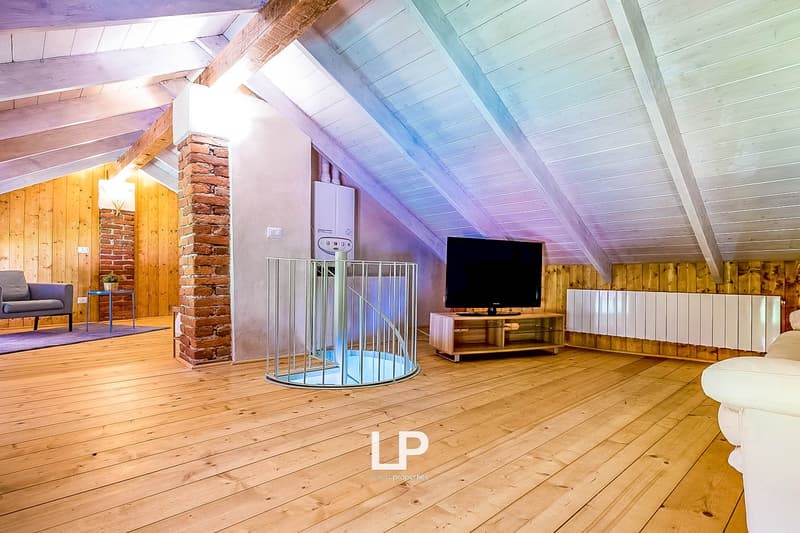 Spacious attic or loft space with wooden floors, exposed wooden beams, and a brick fireplace. The room has a TV stand and seating area, as well as a metal cage-like structure, possibly for storage or decor.