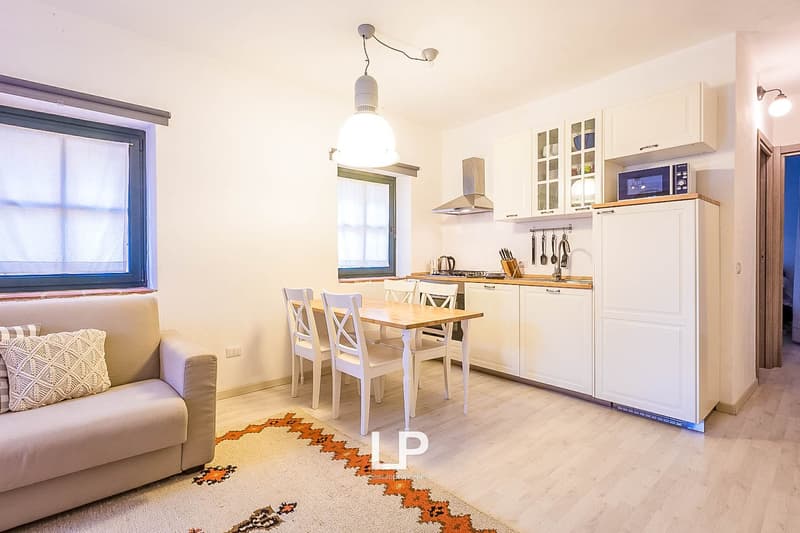 The image shows an open-plan kitchen and living room area. The kitchen features white cabinets, a stainless steel range hood, and a dishwasher. There is a dining table with four chairs in the center of the room. The living room area has a beige sofa and a