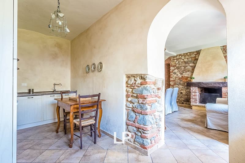 The image shows an open-plan living room and kitchen area. The kitchen features wooden cabinets and a dining table with chairs. There is a stone fireplace visible in the living room area, and the walls have a textured, plaster-like finish. The flooring is