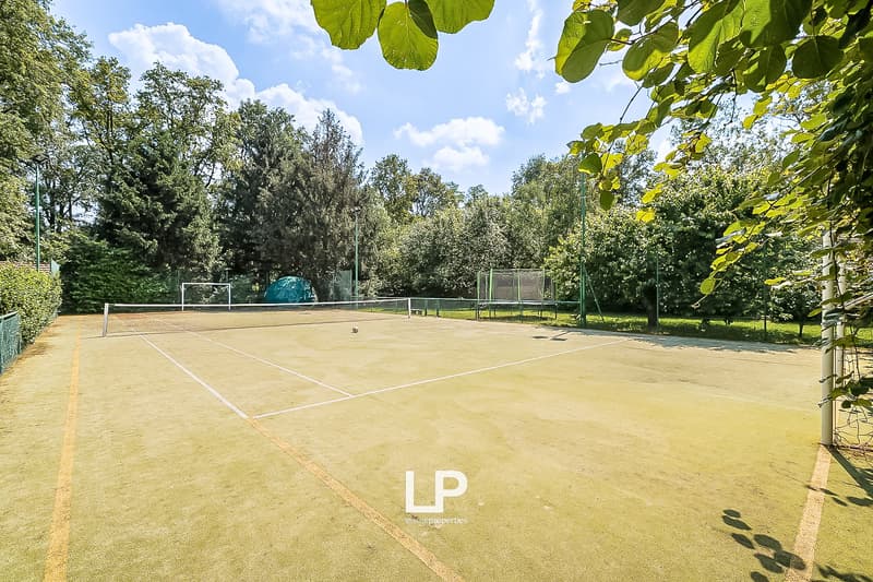 The image shows a tennis court surrounded by lush greenery, including trees and foliage. The court has a clay surface and is enclosed by fencing. There is a green tent or canopy visible on the court.