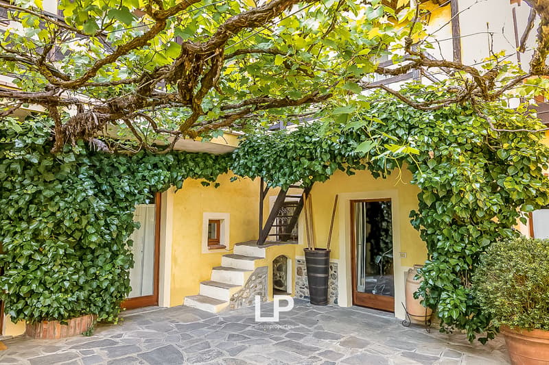 Two-story house with yellow exterior walls, stone steps leading to the entrance, large windows, and a covered patio area surrounded by lush greenery and vines growing on the walls.
