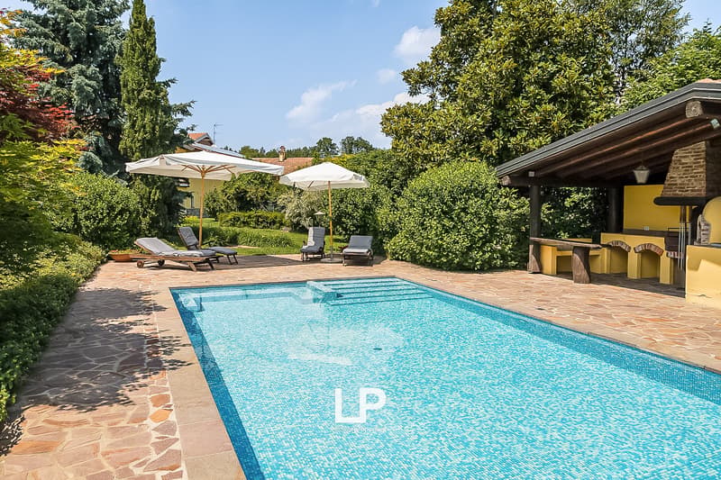 The image shows a beautiful outdoor area with a large swimming pool surrounded by lush greenery and trees. There are sun loungers and umbrellas around the pool, as well as an outdoor kitchen or grill area with a covered patio. The property appears to be a