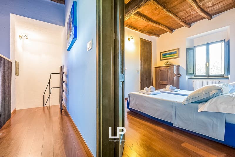 Spacious bedroom with wooden ceiling beams, hardwood floors, and a large window. The room features a double bed with blue bedding, a wooden armoire, and a painting on the wall.