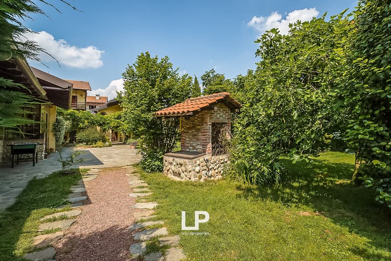 Two-story house with red tile roof, surrounded by lush greenery and trees, stone pathway leading to a small stone structure, patio or terrace area