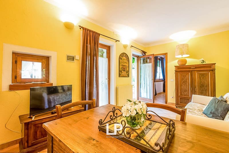 Bright and cozy living room with yellow walls, wooden furniture including a dining table with a decorative centerpiece, a TV stand, and a wooden cabinet. Large windows provide natural light and access to a balcony or terrace.