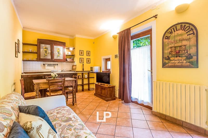 Cozy and bright open-plan living room and kitchen with yellow walls, wooden furniture, tiled floor, and a large window leading to a balcony or terrace. The room features a sofa, a dining table, and a kitchen area with cabinets and appliances. There is als