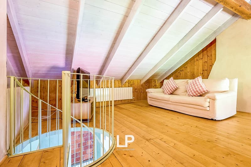 Spacious attic or loft space with wooden beams and paneled ceiling, hardwood flooring, a couch with patterned pillows, and a staircase leading up to the space.