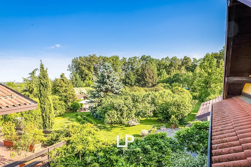 Two-story house with a tiled roof, surrounded by a lush, green garden with various trees and shrubs. The image shows a balcony or terrace overlooking the scenic garden view.