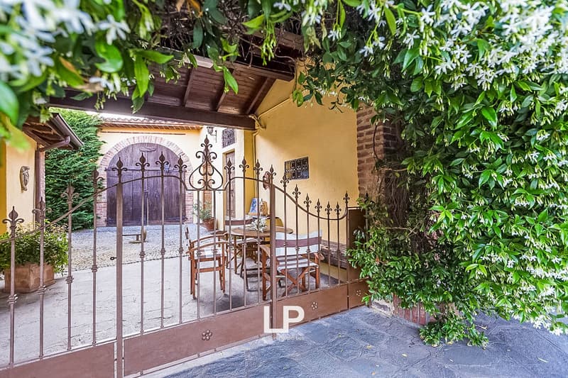 Charming outdoor seating area with wrought iron gates, yellow stucco walls, and a tiled roof. The area is surrounded by lush greenery and flowers, creating a cozy and inviting atmosphere.