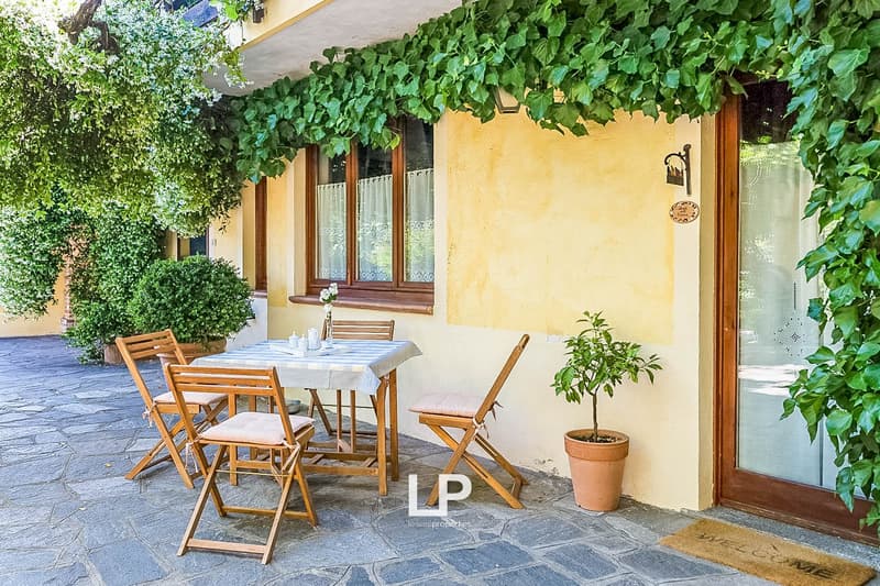Outdoor dining area with wooden table and chairs, surrounded by lush greenery and a yellow stucco wall. The space has a cozy, rustic feel with a potted plant and a covered patio area.