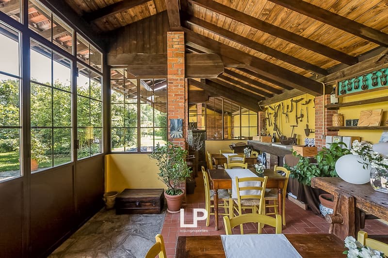 Rustic wooden interior with exposed beams, large windows overlooking a garden, wooden tables and chairs, brick fireplace, and various decorative items and plants throughout the space.