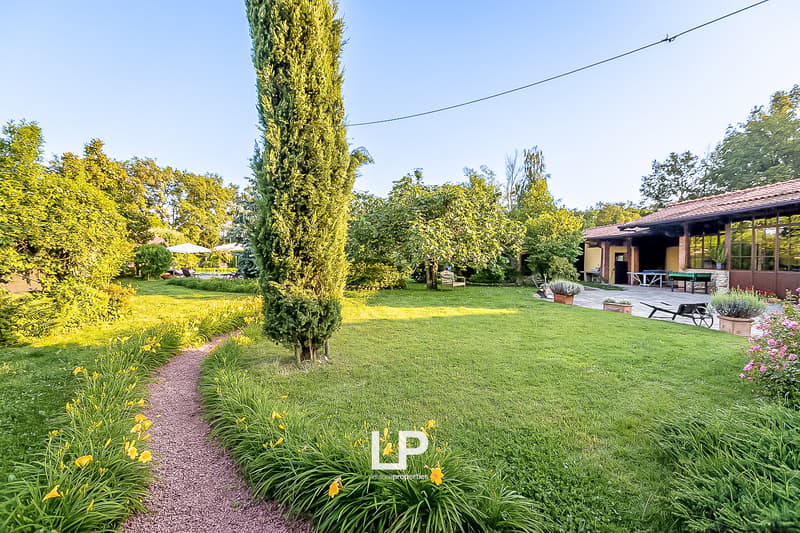 Lush green lawn, colorful flower beds, tall cypress tree, paved walkway, outdoor seating area, and a building with large windows in the background