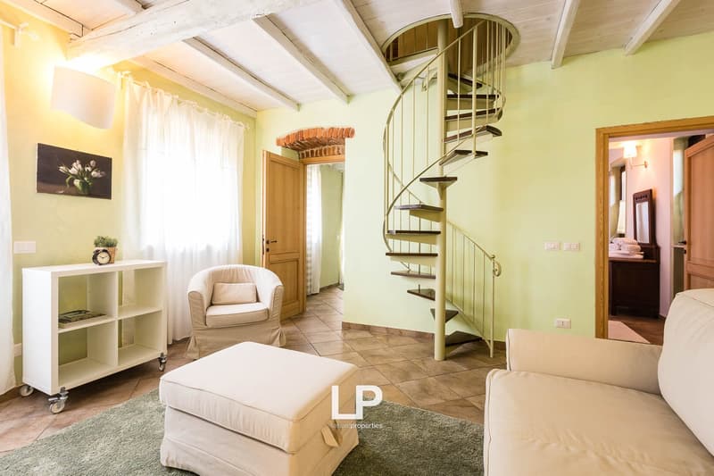 Cozy living room with spiral staircase, wooden furniture, and tiled flooring. The room has a warm, inviting atmosphere with natural light coming through the windows.
