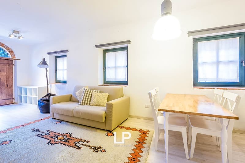 Cozy living room with a yellow sofa, wooden dining table, and large windows providing natural light. The room features a patterned rug and a wooden cabinet.