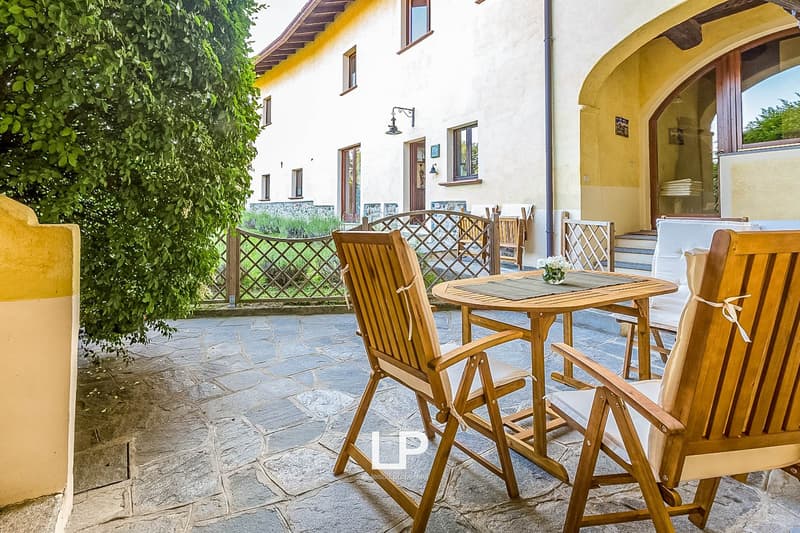 Outdoor patio with wooden table and chairs, stone flooring, arched entryway, and lush greenery surrounding the area.