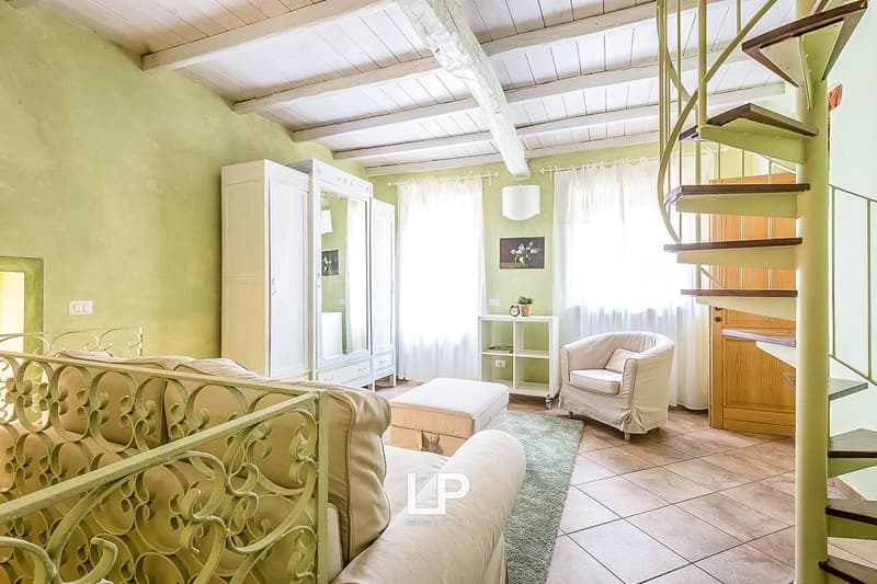 Spacious bedroom with white walls, wooden beamed ceiling, ornate metal bed frame, armchair, shelving unit, and large windows providing natural light
