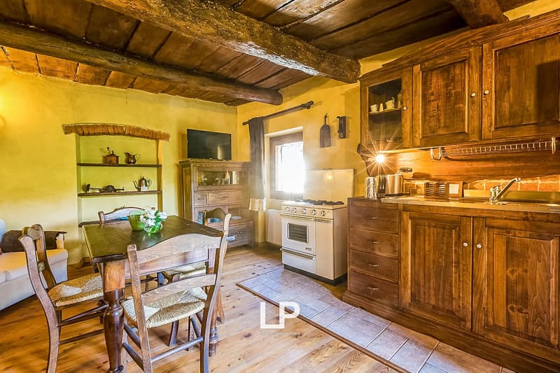 Rustic kitchen with exposed wooden beams, wooden cabinets and shelves, a gas stove, and a dining table with wooden chairs. The room has a warm, cozy atmosphere with a fireplace visible.