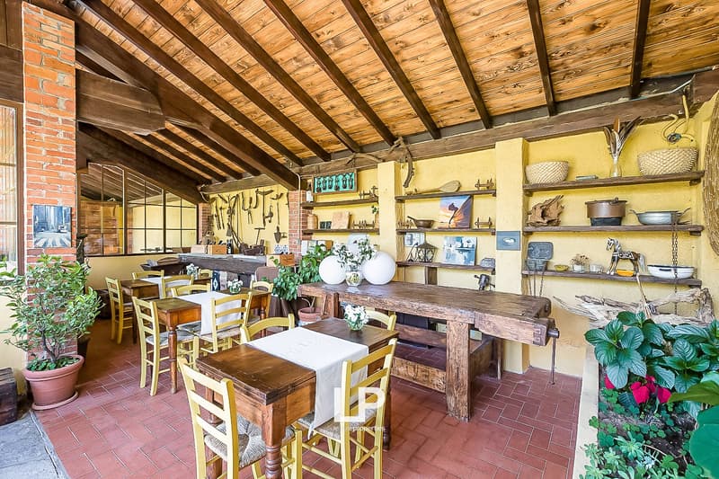 Rustic dining area with wooden beamed ceiling, brick fireplace, wooden tables and chairs, shelves displaying decorative items, and potted plants.