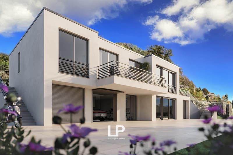 Two-story modern house with a flat roof, large windows, and balconies. The exterior is painted in a light neutral color. There is a garage door visible on the ground floor. The house is surrounded by lush greenery and purple flowers in the foreground.